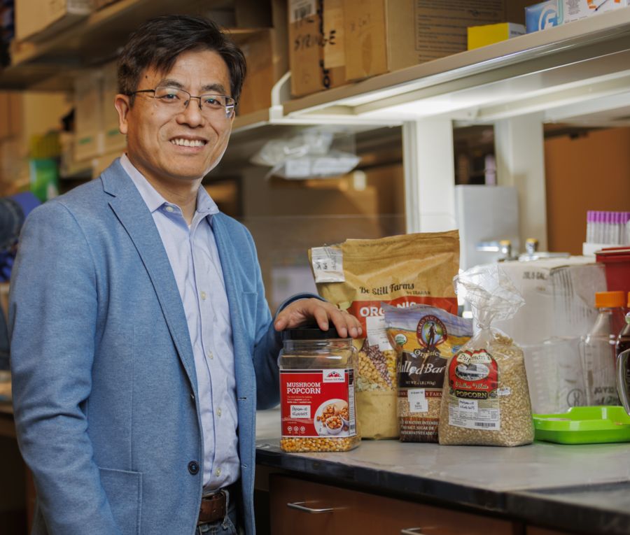 A smiling man in a blue blazer stands beside various containers of popcorn and grains on a countertop, set in a laboratory-like environment filled with shelves and boxes in the background.