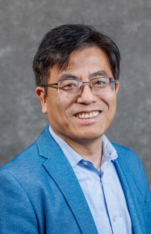 A smiling man with glasses and short black hair poses against a gray backdrop. He is wearing a light blue shirt and a darker blue blazer, exuding a professional and approachable demeanor.