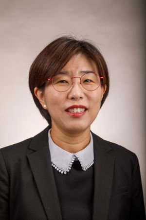 A woman with short hair and red-framed glasses smiles warmly. She wears a black jacket over a blouse with a decorative collar, set against a neutral background.