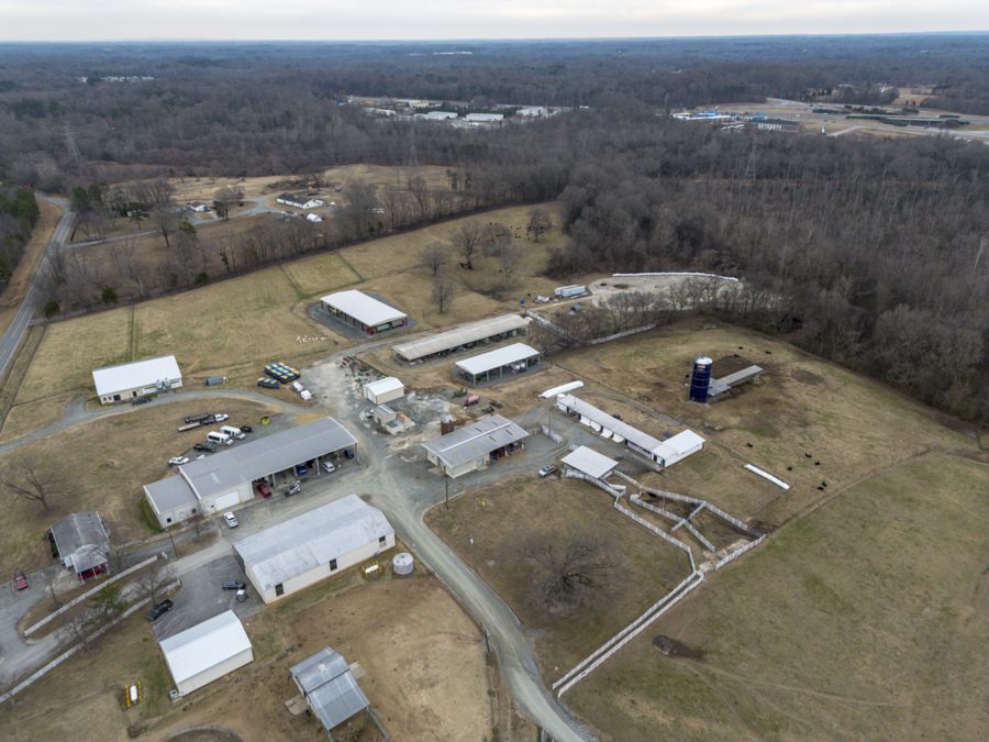 Aerial view of a rural area featuring several buildings, vehicles, and open fields. The landscape includes a mix of structures, including a silos, and several dirt paths connecting different sections of the property, set against a backdrop…