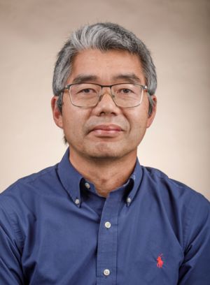 A middle-aged man with gray hair and glasses, wearing a blue button-up shirt. He has a neutral expression and is facing the camera against a light background.