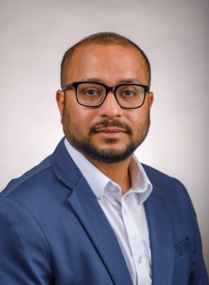 A man with short, dark hair and a beard wears glasses and a blue blazer over a white shirt. He is facing forward with a neutral expression, set against a light background.
