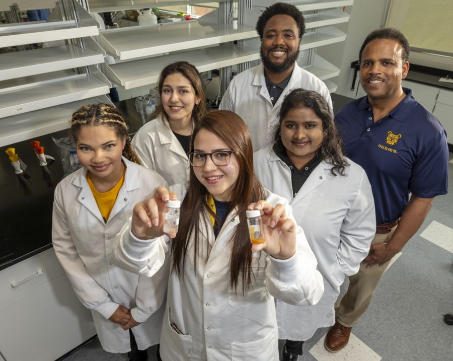 A group of five people in lab coats poses in a laboratory setting. The person in front holds a vial, while others smile behind them. The background features laboratory equipment and shelves.