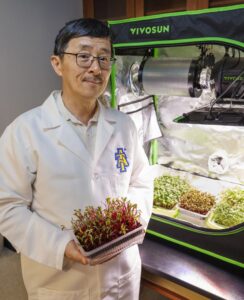 A scientist in a lab coat holds a tray of vibrant red and green microgreens. Behind him is a grow tent with indoor plants, including various types of seedlings under bright lighting.
