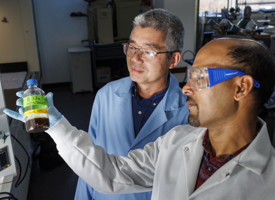 Two scientists in lab coats examine a bottle of brown liquid. One holds the bottle, while the other is closely observing it. Both are wearing safety glasses, and the laboratory setting features equipment in the background.