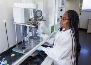 A researcher with long braids works in a laboratory, adjusting a piece of equipment behind a safety glass enclosure. She wears protective gloves and a lab coat, focusing intently on her task.