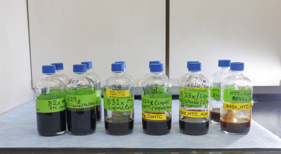 A lineup of glass bottles filled with various colored liquids, each labeled with bright green and yellow tags indicating different sample names and dates. The bottles are arranged on a surface, with some filled with dark liquids and others…