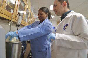 Two researchers in lab coats collaborate in a laboratory setting. One person, wearing a blue lab coat and gloves, works with a mixer, while the other, in a white lab coat, observes attentively. Cabinets filled with equipment line the…