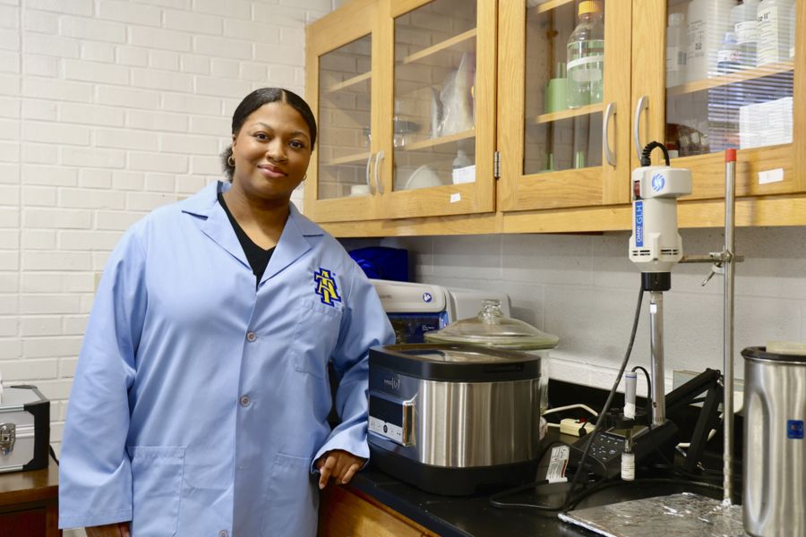 A woman in a light blue lab coat stands in a laboratory, smiling at the camera. She is next to a countertop with scientific equipment and various containers in wooden cabinets behind her.