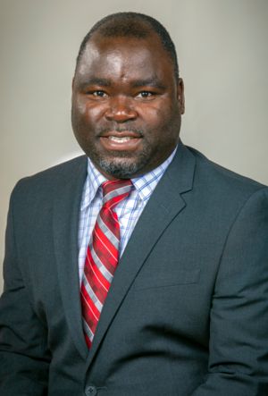 A professional man smiles at the camera, wearing a dark suit and a checked shirt with a red and white striped tie. He has a short beard and stands against a neutral background.