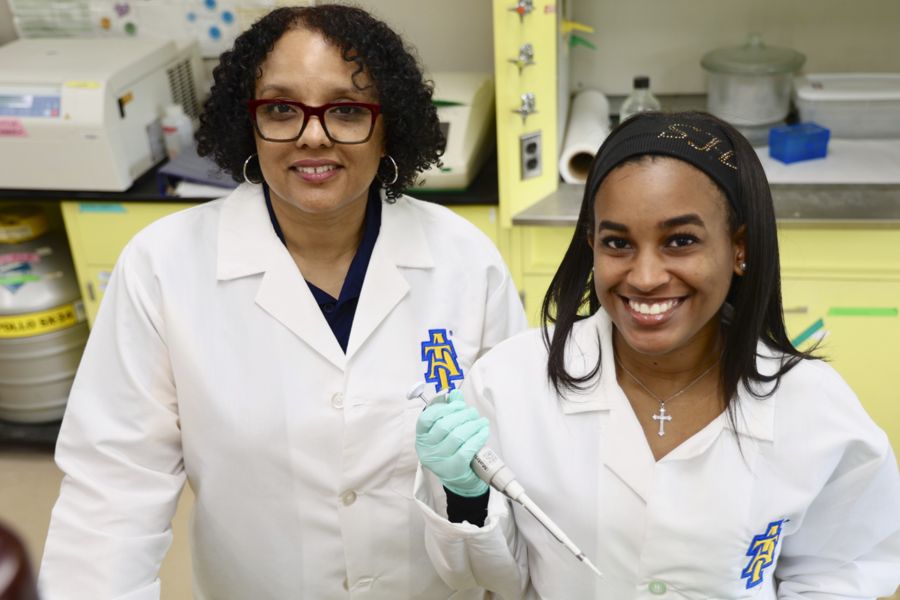 Two women in lab coats smile in a laboratory setting. One holds a pipette with a teal glove, while the other looks directly at the camera. Lab equipment and supplies are visible in the background.