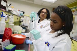 A young woman in a lab coat carefully uses a micropipette to measure a substance in a laboratory, while another woman in the background observes. Various lab equipment and materials like colored containers are visible on the countertop.