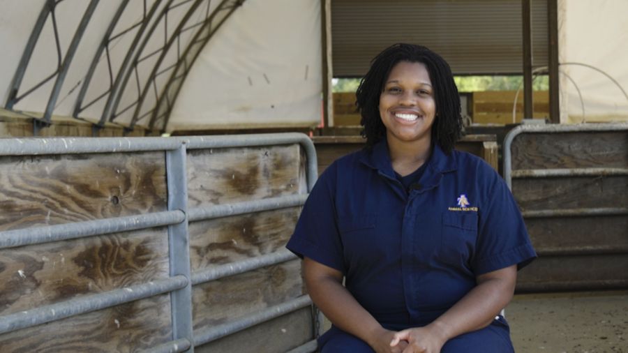 A woman with curly hair smiles while sitting on a bench in a barn-like structure. She is dressed in a blue uniform, surrounded by wooden fencing. Natural light illuminates the scene, creating a welcoming atmosphere.
