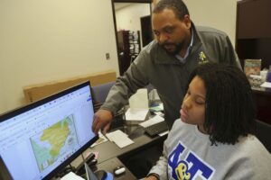 A man gestures toward a computer screen displaying a map while a woman sits at the desk, focused on her work. The office environment features paperwork and various office supplies.
