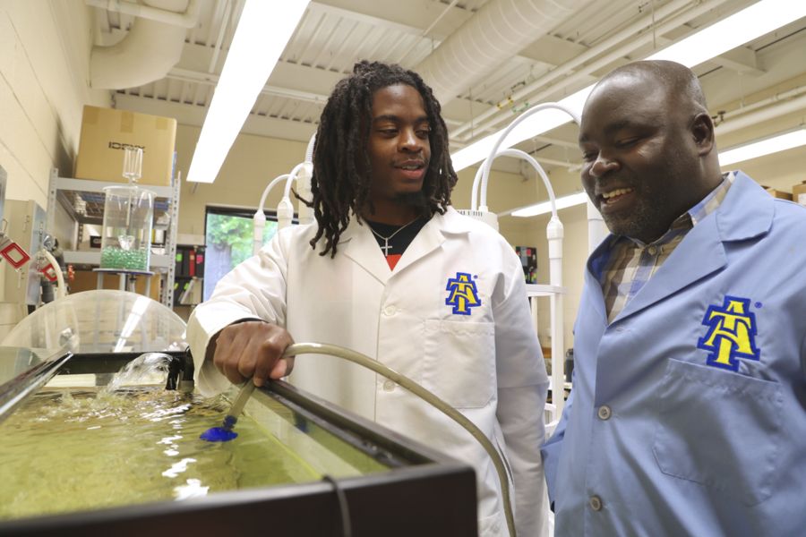 Two individuals in lab coats interact by a fish tank in a laboratory. One person uses a tube to manage water while the other smiles, suggesting a collaborative atmosphere in a scientific setting.