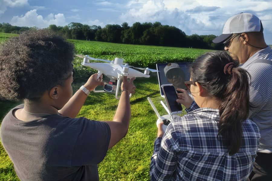 A group of three individuals is outdoors near a green field, focused on flying a drone. One person is holding the drone while another operates a controller, and a third is looking at a device displaying information.