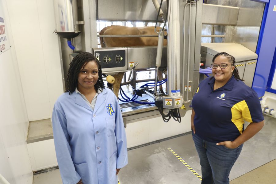 Two women stand in a dairy facility, one wearing a lab coat and the other a blue and yellow polo shirt. Behind them is a milking machine with a cow partially visible. The environment appears clean and professional.